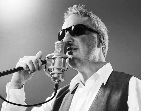 Ein Mann mit Sonnenbrille singt in ein Mikrofon. Er trägt eine Weste über einem weißen Hemd und hat graue Haare. Der Hintergrund ist neutral und unscharf, was den Fokus auf den Sänger lenkt.