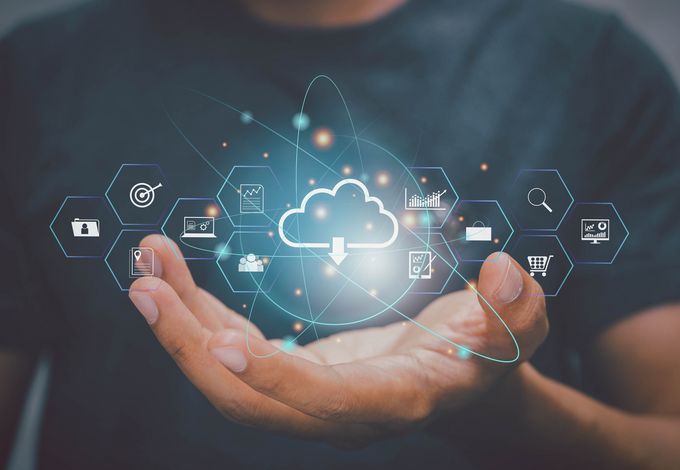 Eine Hand hält ein leuchtendes Cloud-Symbol, umgeben von verschiedenen Icons, die digitale Technologien und Daten darstellen, wie Grafiken, Einkaufskörbe und Sicherheitszeichen. Der Hintergrund ist unscharf und zeigt eine Person in einem dunklen Shirt.