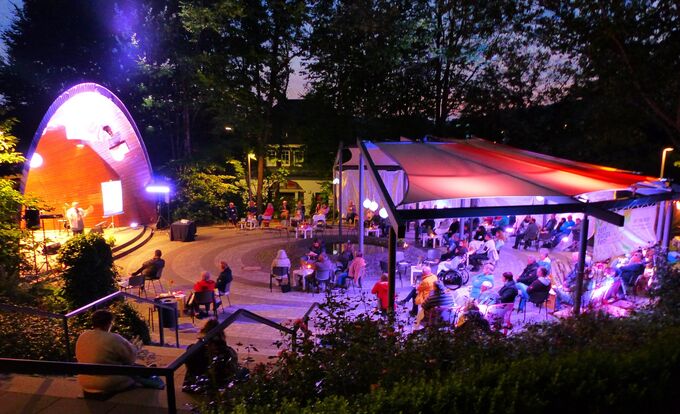 Eine Außenveranstaltung bei Nacht mit einer Bühne, auf der ein Musiker auftritt. Im Vordergrund sitzen viele Menschen an Tischen unter einem überdachten Bereich. Bunte Lichter erhellen die Szene und schaffen eine festliche Atmosphäre.