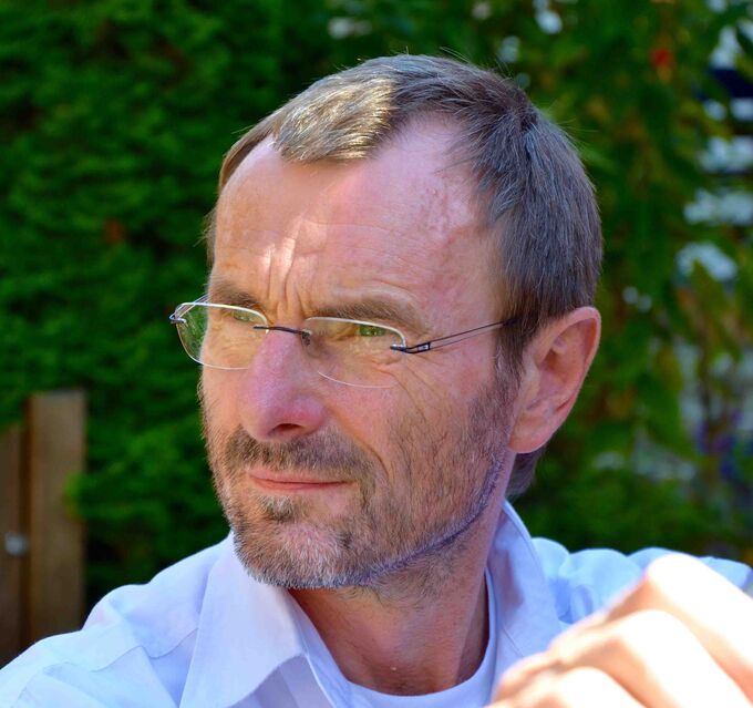 Ein Mann mit Brille und kurzem, grauem Haar schaut nachdenklich in die Ferne. Er trägt ein helles Hemd und steht vor einer grünen, bewachsenen Kulisse. (Text KI generiert)
