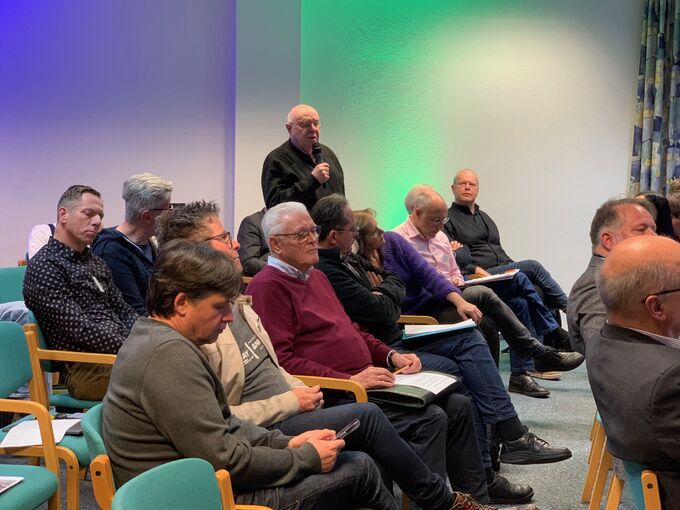 Eine Gruppe von Männern sitzt in einem Raum, einige hören zu, während ein Mann im Vordergrund spricht. Der Raum hat eine grüne und blaue Beleuchtung. Die Teilnehmer scheinen an einer Diskussion oder Versammlung teilzunehmen.