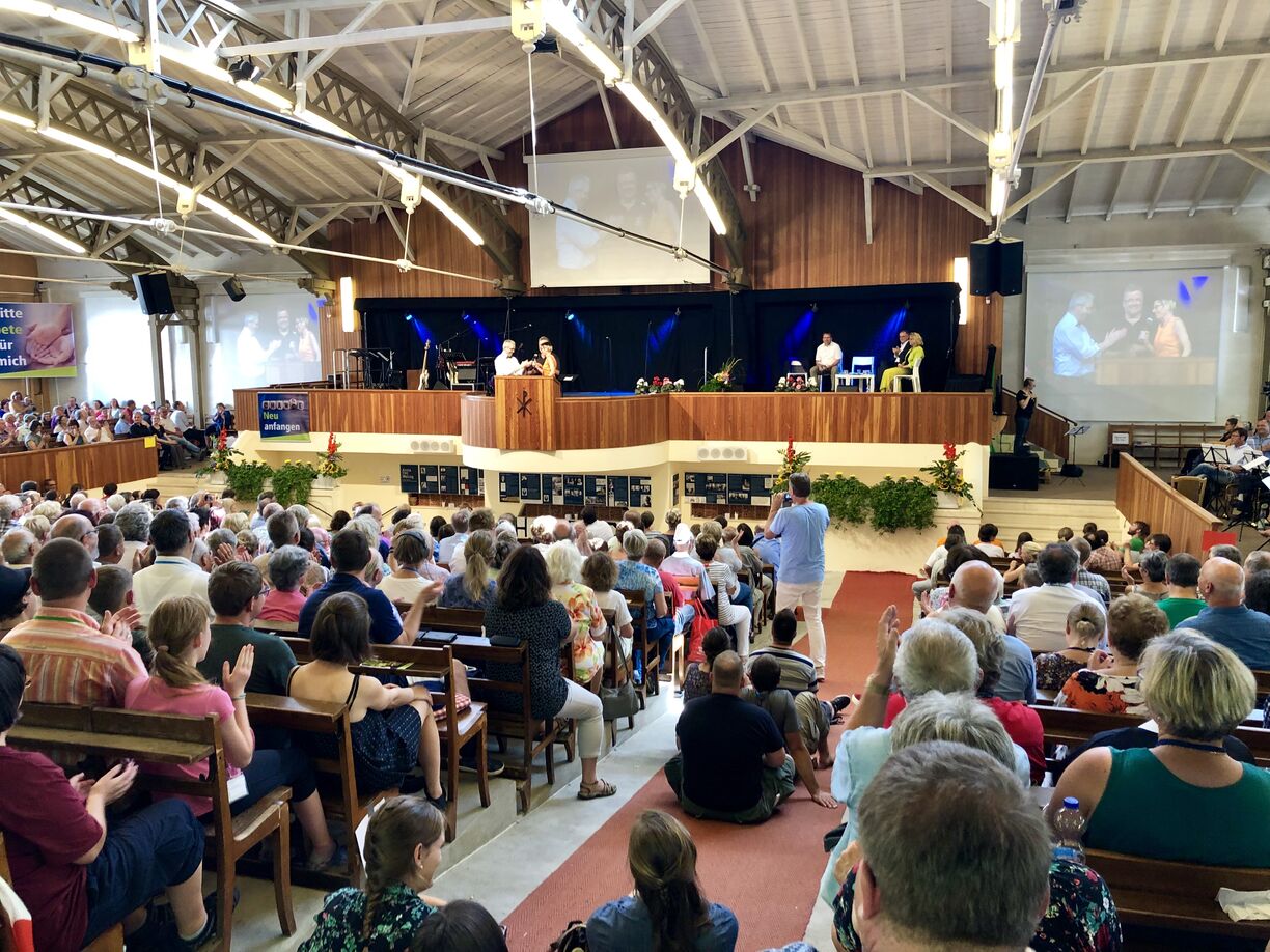 Eine große Menschenmenge sitzt in einem hellen, modernen Veranstaltungsraum. Auf einer Bühne steht ein Redner, während andere Personen auf Stühlen sitzen. Die Zuschauer applaudieren, und im Hintergrund sind Lichter und eine Leinwand sichtbar.