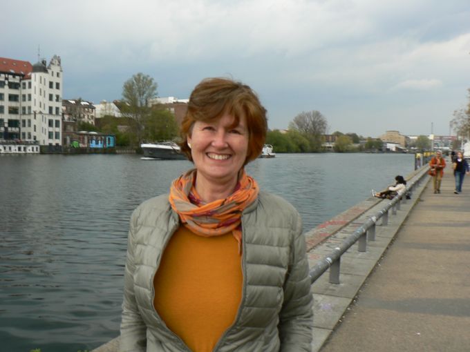 Eine lächelnde Frau mit braunen Haaren steht am Ufer eines Flusses. Sie trägt eine graue Jacke und einen orangefarbenen Pullover sowie einen bunten Schal. Im Hintergrund sind Bäume und Gebäude zu sehen, während einige Personen am Wasser sitzen.