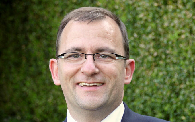 Ein Mann mit Brille lächelt in die Kamera. Er trägt einen dunklen Anzug, ein weißes Hemd und eine rote Krawatte mit Punkten. Im Hintergrund sind grüne Pflanzen sichtbar.