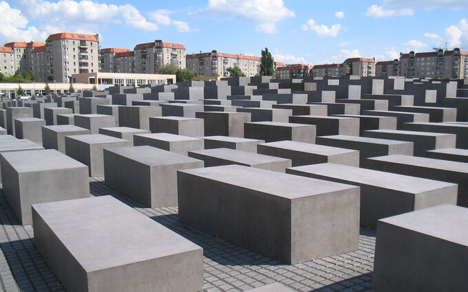Das Bild zeigt das Holocaust-Mahnmal in Berlin, bestehend aus vielen grauen Betonblöcken, die in einem unregelmäßigen Muster angeordnet sind. Im Hintergrund sind Wohngebäude und ein blauer Himmel mit Wolken sichtbar.