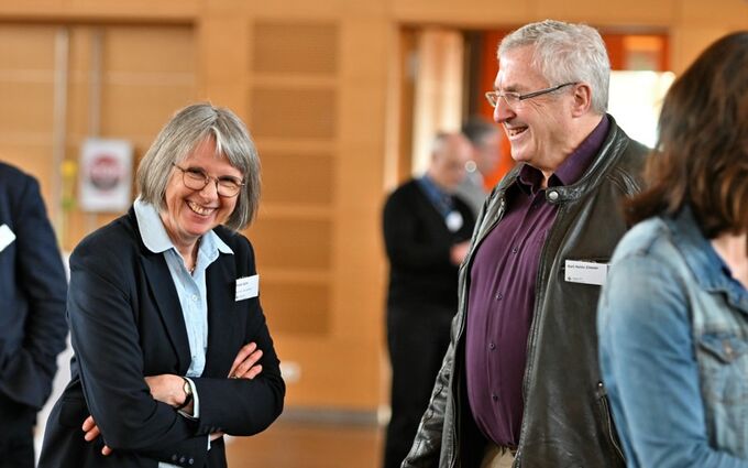 Zwei Personen stehen in einem Raum und lachen miteinander. Eine Frau mit Brille und grauen Haaren trägt eine dunkle Jacke, während ein älterer Mann in einer Lederjacke ebenfalls lächelt. Im Hintergrund sind weitere Personen zu sehen.
