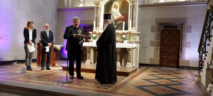 Eine Zeremonie in einem Kirchenraum. Ein Mann in schwarzer Robe spricht zu einem anderen Mann, der eine ähnliche Kleidung trägt. Im Hintergrund stehen zwei Personen, während ein Altar mit einer religiösen Darstellung sichtbar ist. Die Beleuchtung ist sanft und es gibt dekorative Elemente im Raum.