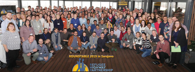 Gruppenfoto von über 200 Teilnehmern eines Roundtable-Events in Sarajevo, 2019. Die Menschen stehen in mehreren Reihen, lächeln und tragen unterschiedliche Kleidung. Im Vordergrund ist ein Banner mit dem Schriftzug "Refugee Highway Partnership" zu sehen.