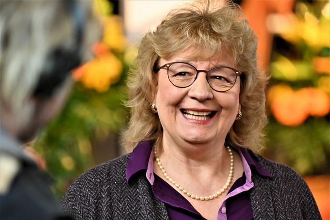 Eine lächelnde Frau mit blonden, lockigen Haaren und Brille trägt eine Perlenkette und ein lila Hemd unter einem grauen Blazer. Sie steht vor einer bunten Blumenarrangement und scheint mit jemandem zu sprechen.