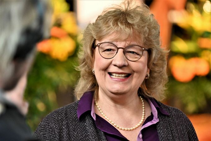 Eine lächelnde Frau mit lockigem, blonden Haar und Brille trägt eine Perlenkette und ein lila Oberteil. Sie steht vor einer bunten, blühenden Kulisse und scheint mit jemandem zu sprechen.
