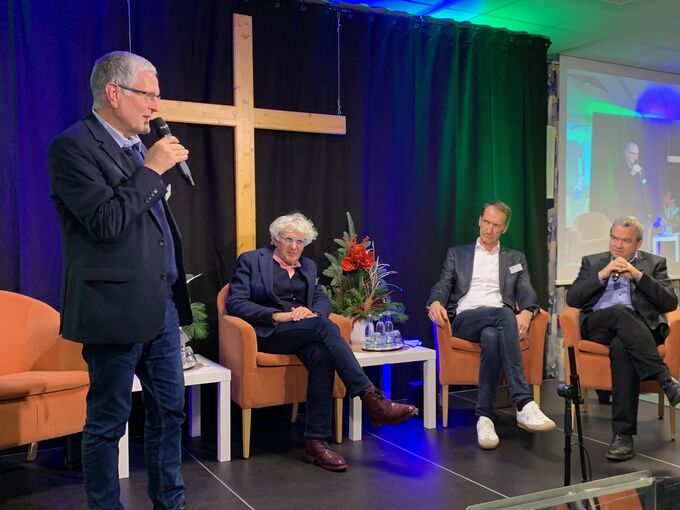 Eine Diskussionsrunde auf einer Bühne mit vier Personen. Ein Mann spricht ins Mikrofon, während die anderen drei auf Sesseln sitzen. Im Hintergrund ist ein Kreuz und eine dekorative Pflanzenanordnung zu sehen.