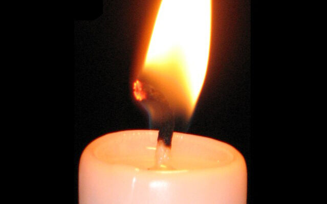 Eine brennende weiße Kerze auf schwarzem Hintergrund. Die Flamme leuchtet hell und erzeugt eine warme Atmosphäre.