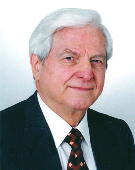 Ein älterer Mann mit grauen Haaren und einem freundlichen Gesichtsausdruck trägt einen dunklen Anzug mit einer gestreiften Krawatte. Der Hintergrund ist hell und neutral.